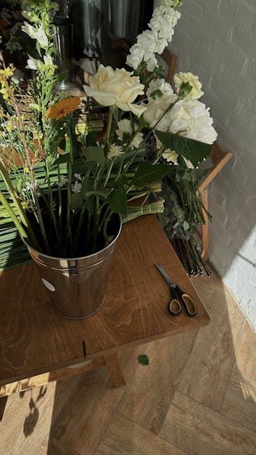 A tall, elegant bouquet featuring large white roses, white carnations, and smaller white filler flowers arranged with green foliage, all presented in a shiny metallic bucket. The bouquet is placed on 