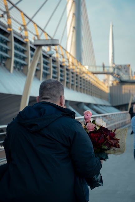 A man standing outdoors near a bridge, holding a bouquet of fresh flowers wrapped in light brown paper. The bouquet features a mix of white, pink, and red blooms, including roses and other floral vari