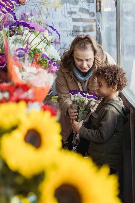 A woman and a young boy are inside a flower shop, engaging in a flower delivery transaction. The woman, with curly blonde hair and a beige coat, gently hands a small bouquet of purple and white flower