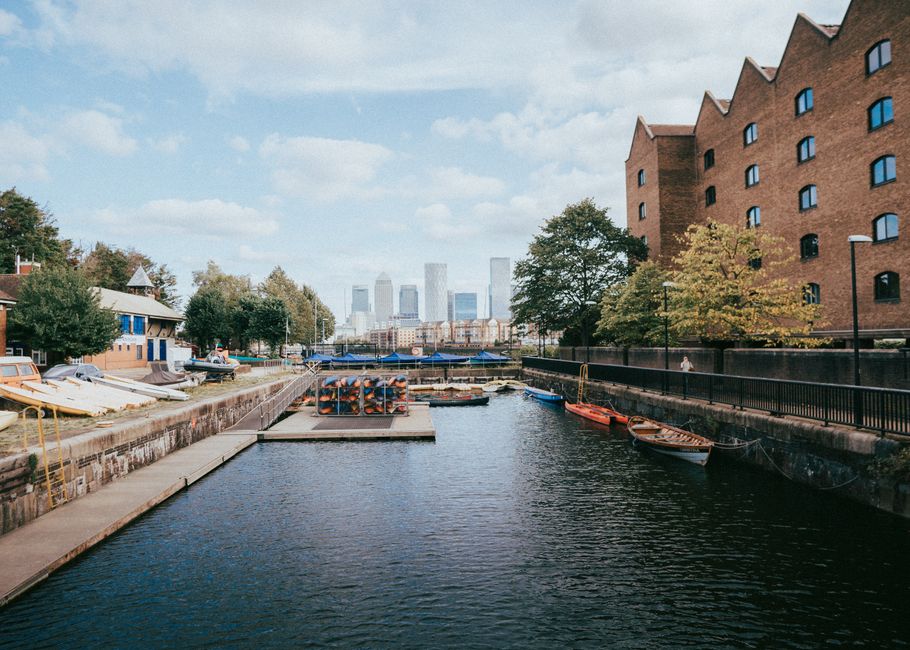 The image depicts a calm waterway bordered by a paved walkway on the left side, with various small boats moored along the edge, including some with orange and red hulls. On the right, there is a large