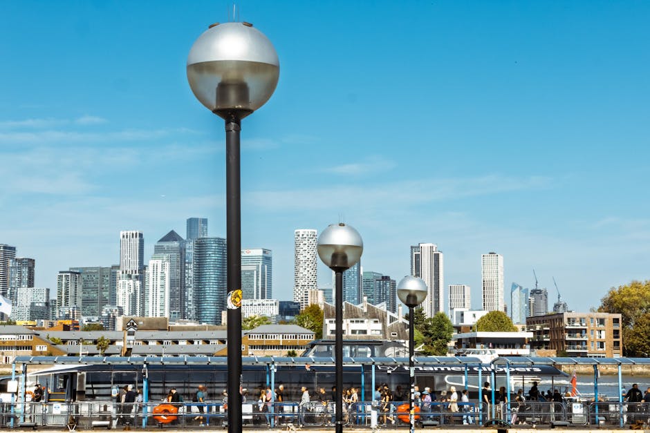 The image depicts a cityscape with modern skyscrapers and office buildings in the background under a clear blue sky. In the foreground, there are five evenly spaced street lamps with spherical, metall