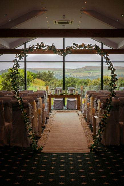 A circular floral arch decorated with lush green foliage and assorted flowers surrounds a wedding aisle in a modern venue with large panoramic windows revealing a scenic countryside view. The aisle is