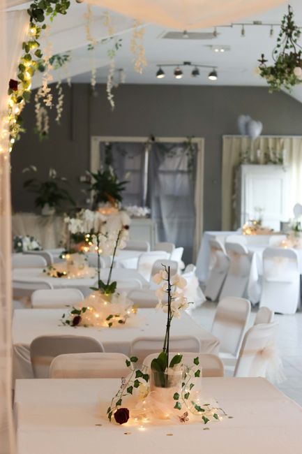A floral arrangement featuring white orchids and dark red roses placed in a metallic vase on a table, surrounded by delicate string lights and scattered flower petals. The setting is an elegant indoor