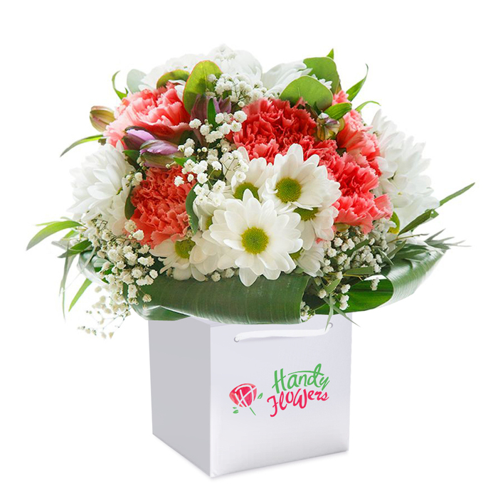 Captured against a flawless white background, this sensational Leamouth bouquet showcases the artistry and freshness local florists take pride in. An upright white square gift bag, fitted with elegant ribbon handles, serves as the contemporary vessel for a lively array of seasonal blooms. The focal point comprises a sumptuous mix of coral and soft peach carnations, each flower's frilled petals lending depth and tactile beauty, their tones matched by pops of vibrant red. Scattered in between are bright white daisy-like blossoms with chartreuse centers, recalling the cheerful vibrancy of local markets on Leamouth Road. Sprigs of baby's breath, or gypsophila, contribute a gossamer lightness, their mini blooms softly diffused throughout the display. Framing the entire arrangement are broad, glossy deep-green leaves and additional lush foliage, evocative of the greenery seen along Bow Creek Ecology Park. The composition is made all the more inviting by the emergence of several purple-tinged buds, slightly closed, peeking from the left side and adding hidden layers to the whole. The artistry of the bouquet lies not only in its selection but in its harmonious, dome-like shape, exuding a sense of abundance, care, and readiness for gifting. With its crisp packaging and balanced palette, this floral creation is perfect for marking special occasions-be it an anniversary, heartfelt thank you, or joyous celebration. The bouquet stands as a beautiful representation of Leamouth's fresh, contemporary floral scene, available for same-day delivery to homes and offices across the area.