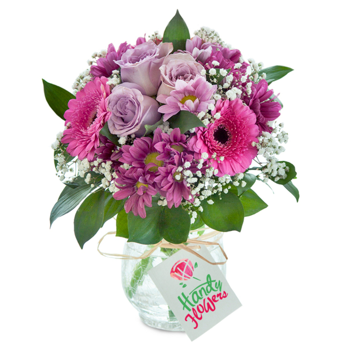At the centre of Leamouth's vibrant flower scene, this meticulously arranged bouquet glows with artistry and charm. Set before a pristine white background, the arrangement is anchored by sumptuous lavender roses gently opening in the middle-each bloom a testament to freshness reminiscent of early morning walks by the Limehouse Cut. Large, hot pink Gerbera daisies, unmistakable with their dark centres and exuberant flair, surround the roses. These are harmonized by clusters of magenta chrysanthemums, some sporting lively greenish cores, which add texture and richness to the display. Sprigs of dainty white baby's breath are woven in, creating a whimsical, cloud-like effect that softens the overall shape and highlights every hue. Deep emerald foliage peeks from beneath, lending both contrast and lush fragrance that fills the air like a breeze off the nearby riverfront. The bouquet rests in a clear glass vase whose unique cracked texture plays with the light and shadow, giving it a tactile rustic beauty. An unfussy raffia bow circles the vase's neck, holding a plain white tag-ready for a heartfelt note for birthdays, thank-you gifts, or surprise deliveries to homes off Goodluck Hope. This arrangement is a fine example of the local florist's craft-thoughtful, inviting, and an ideal gift to add warmth to any occasion in Leamouth.