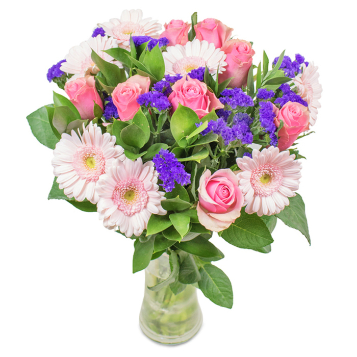 A striking bouquet stands poised in a slender clear vase, its composition echoing the gentle beauty found along the waterside lanes of Leamouth. Approximately seven to nine delicate pink roses, some tightly budded while others just begin to unfurl, present a classic spiraled form that speaks of both romance and new beginnings. Large pale pink gerbera daisies, with luminous centers shading toward near-white, radiate like soft morning sunshine reflecting on the Thames. Dotted between, generous clusters of vibrant purple statice add textured contrast and a playful pop of colour, reminiscent of the wild flora in East India Dock Park. Lush, deep green foliage gathers beneath, providing both structure and a breath of freshness, while the transparent vase-filled with water and crisp visible stems-suggests same-day freshness and expert preparation by local hands. This arrangement, suitable for joyful milestones or a touch of everyday grace in a Leamouth home or office, conveys a spirit of gentle celebration and heartfelt connection rooted in its local artistry.